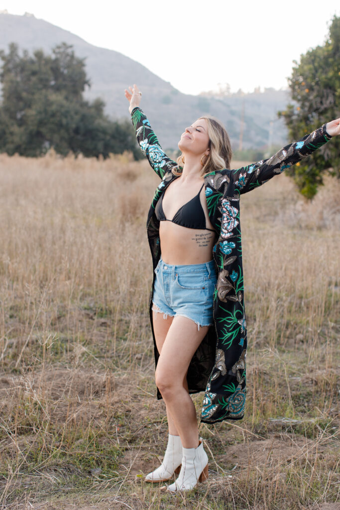 Alessia Citro in a pasture wearing a kimono, bikini top, and cut off shorts. She looks free as a bird, because she is, thanks to habits.